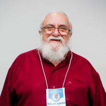 Retrato de Wilson Savino. Cabelo e barba longos e brancos, com uma camisa social vermelha